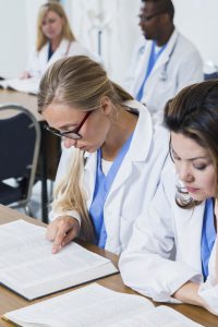 Close up of medical students sitting at a table in a classroom, wearing white lab coats over blue scrubs. The focus is on the blond woman wearing eyeglasses, reading a textbook.